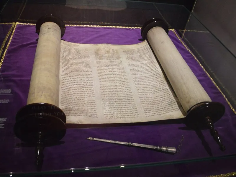 An open Torah scroll beside a printed Bible on a wooden table